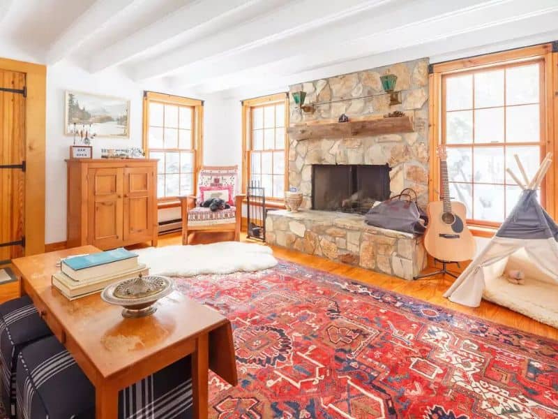 Cozy and warm living room with a stone fireplace, comfortable seating, a large red patterned area rug, and rustic decor in a Catskills rental cabin.