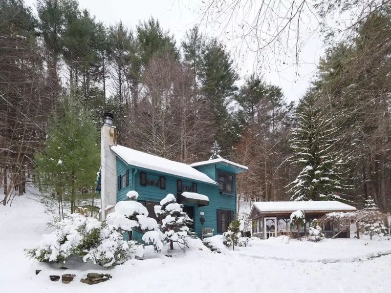 Exterior of a vibrant blue winter Catskills cabin rental blanketed in snow, nestled among tall pine trees.