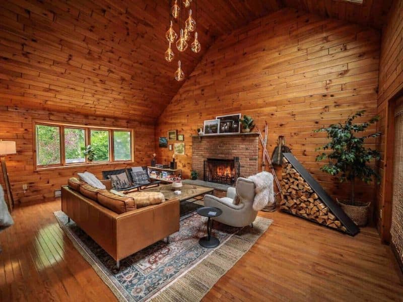 Spacious living room inside a cozy cabin in the Catskills, featuring a brick fireplace with a woodpile, a comfortable sofa, and large windows allowing natural light.