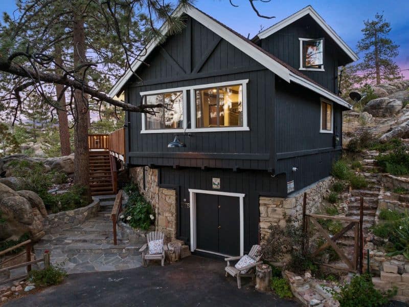 Exterior of a two-story Big Bear Lakefront cabin rental with black siding and a stone foundation, with stairs leading to the front door.