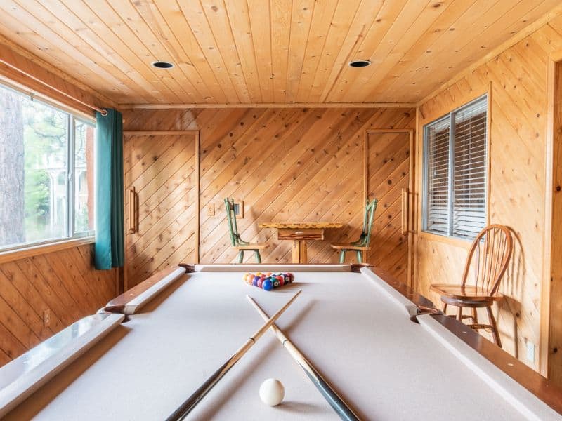 Billiards room in a lakefront cabin featuring a pool table with cues crossed over, wood-paneled walls, and a window with a forest view.
