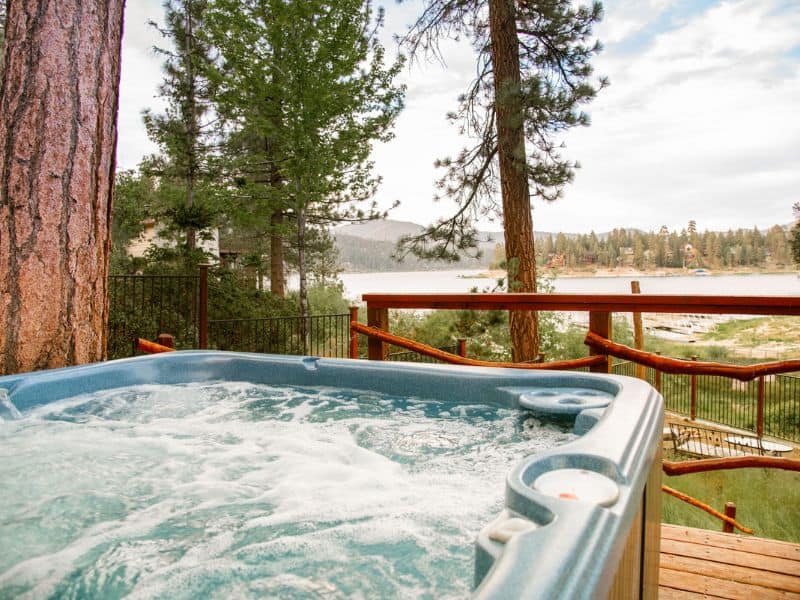 Outdoor hot tub on the deck of a Big Bear lakefront cabin with a railing overlooking the lake, surrounded by tall pine trees.