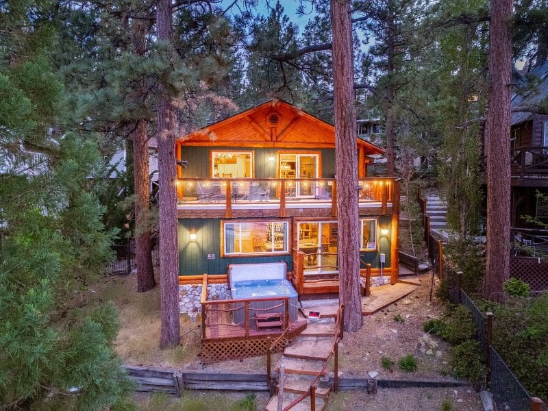 Twilight view of a multi-story Big Bear lakefront cabin rental with warm interior lighting, a deck, and hot tub, surrounded by pine trees.
