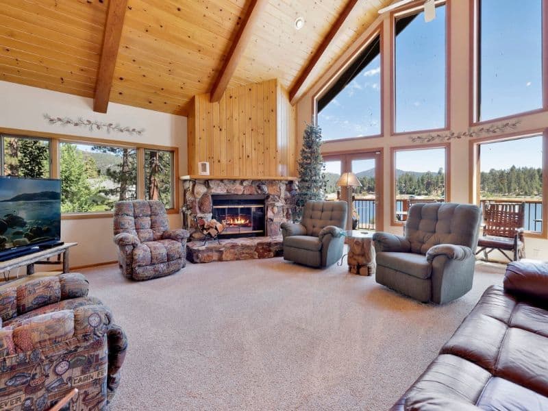 Living room with a stone fireplace, plush seating, and large windows overlooking the lake in a cabin rental in Big Bear.