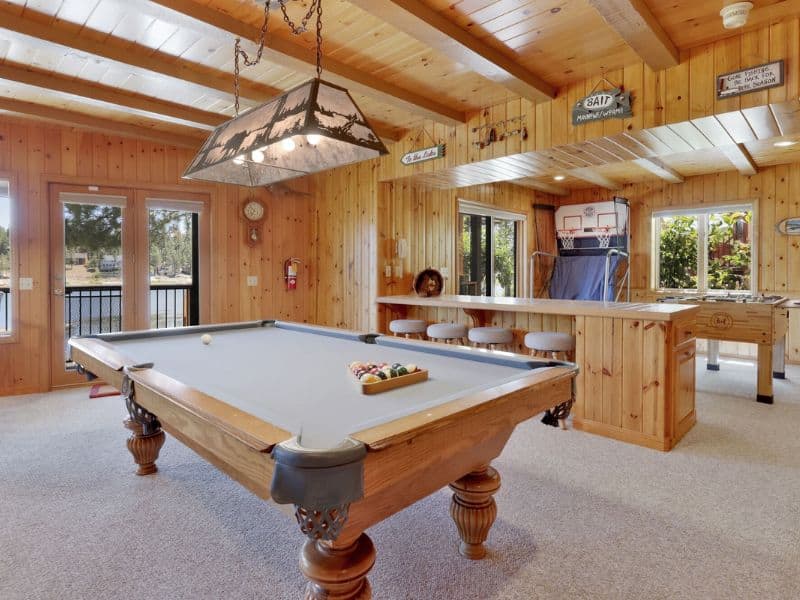 Game room in a Big Bear cabin featuring a pool table, a basketball hoop game, and lake view windows, with wood-paneled walls.