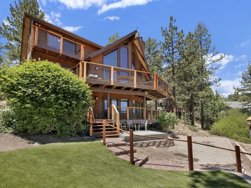 A spacious Big Bear lakefront cabin rental with a large deck and green lawn, set against a backdrop of mature trees.