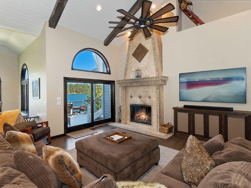 Living room inside a Big Bear rental with vaulted ceilings, a grand fireplace, plush sofas, and double doors opening to a lake-view balcony.