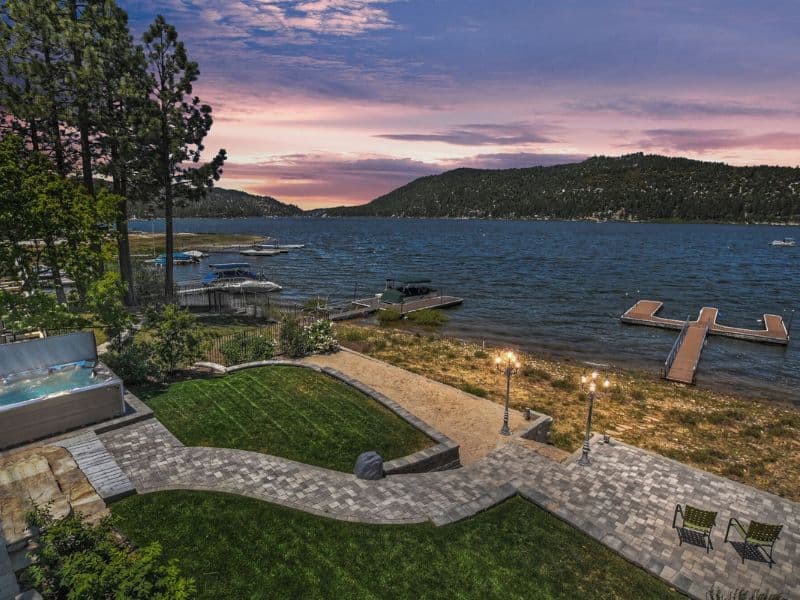 Twilight view of a Big Bear lakefront cabin backyard with lit path leading to a private dock on the lake, as well as outdoor seating and a hot tub.