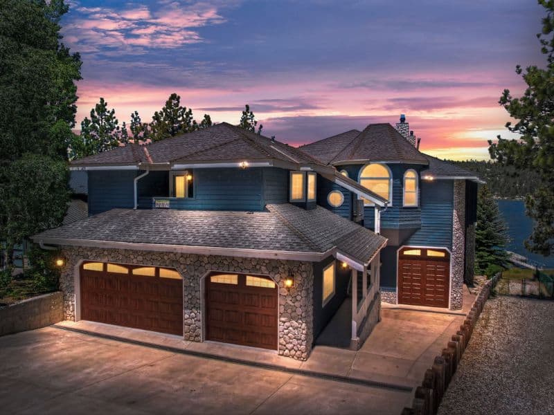 Exterior shot of a multi-story Big Bear Vrbo rental at dusk, showcasing its large windows, stone accents, and a double garage with a driveway.