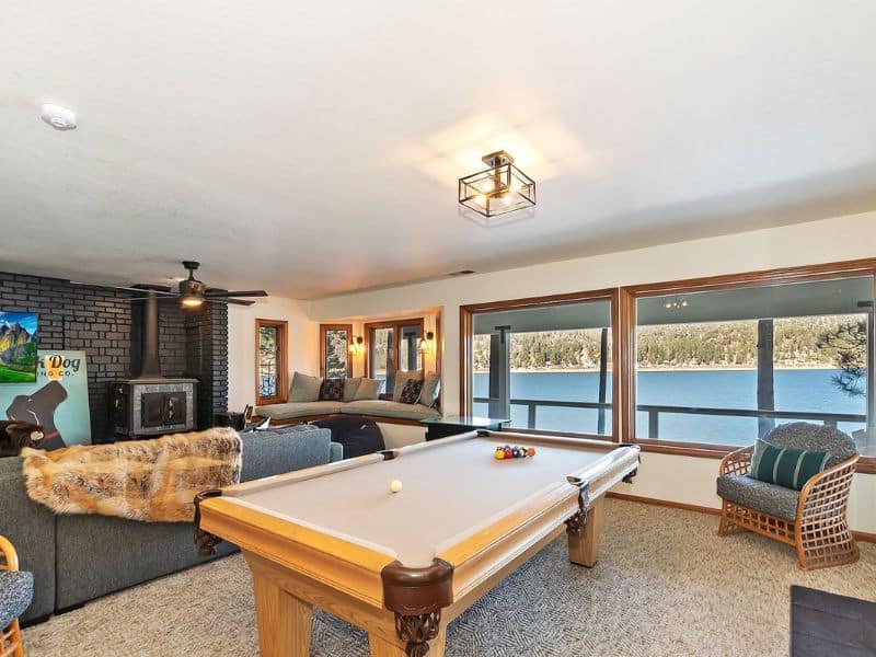 Game room in a lakefront cabin rental in Big Bear, complete with a billiards table, wood stove, and ample seating, framed by large windows with lake views.