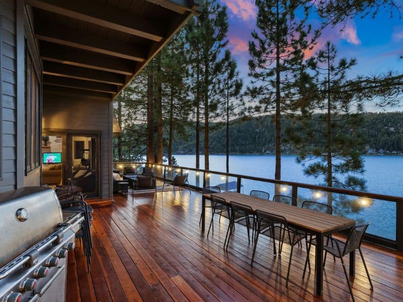Spacious deck of a Big Bear lakefront cabin at dusk, featuring outdoor dining and a grill, with a view of the lake through the pines.