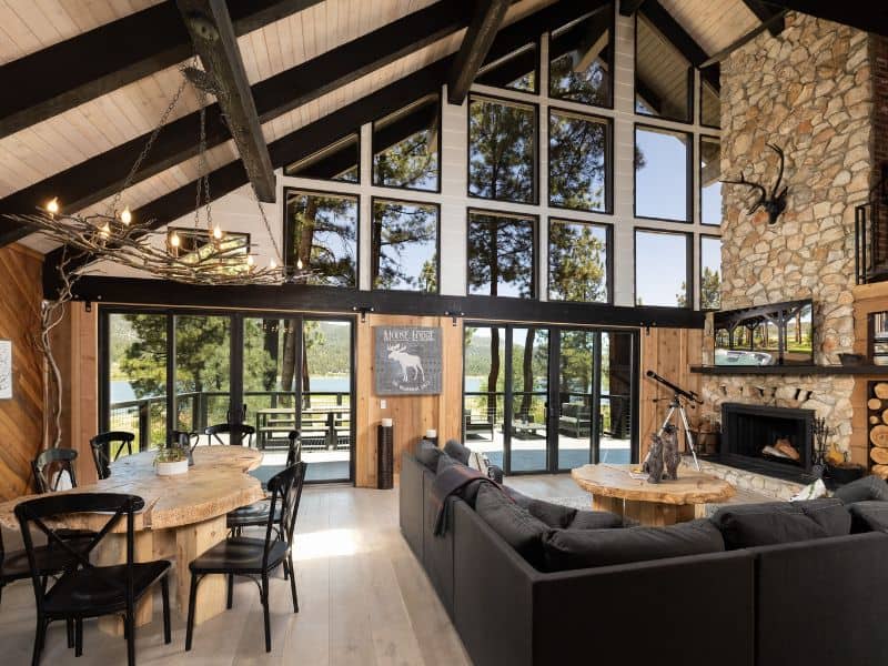 Interior of a Big Bear lakefront cabin with high ceilings, featuring a stone fireplace, a rustic chandelier, and floor-to-ceiling windows offering a view of the surrounding lake and forest.