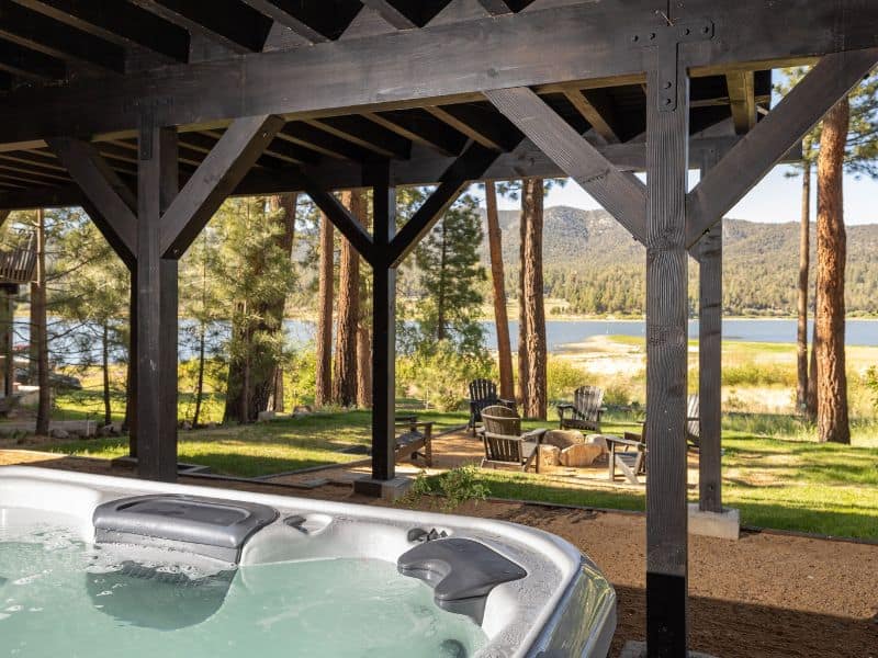 Outdoor hot tub on a covered patio of a lakefront cabin in Big Bear, with Adirondack chairs overlooking the lake and forest.