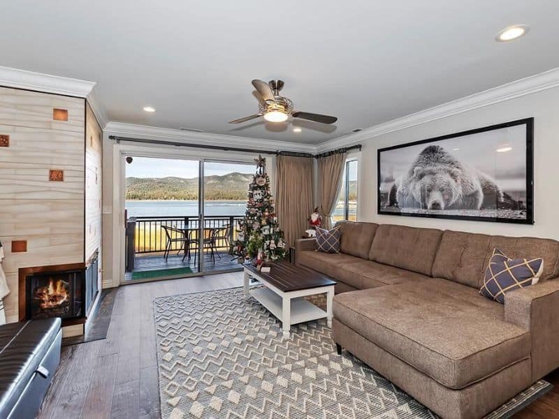 Spacious living room with a fireplace in a Big Bear Lakefront condo rental, featuring comfortable seating and festive holiday decorations with lake views through the balcony door.