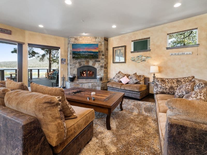 Comfortable living room in a Big Bear rental with a large fireplace, plush sofas, and windows revealing a lake view.