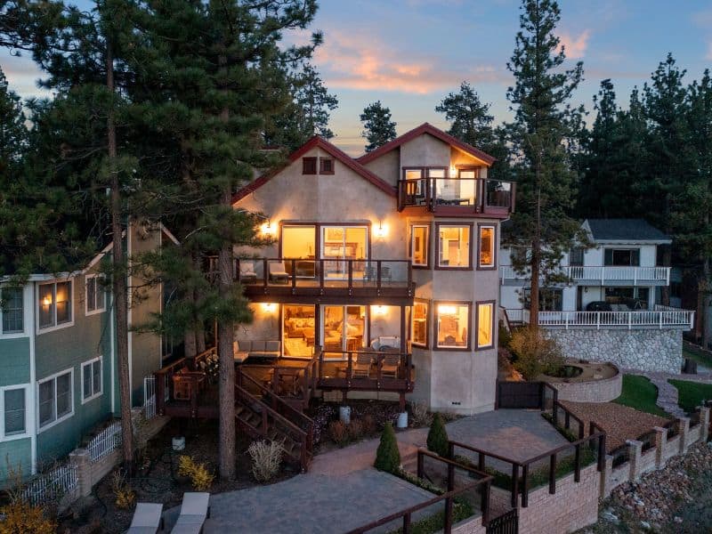 Evening view of a multi-level Big Bear Vrbo rental with illuminated windows, balconies overlooking the lake, and a landscaped garden.