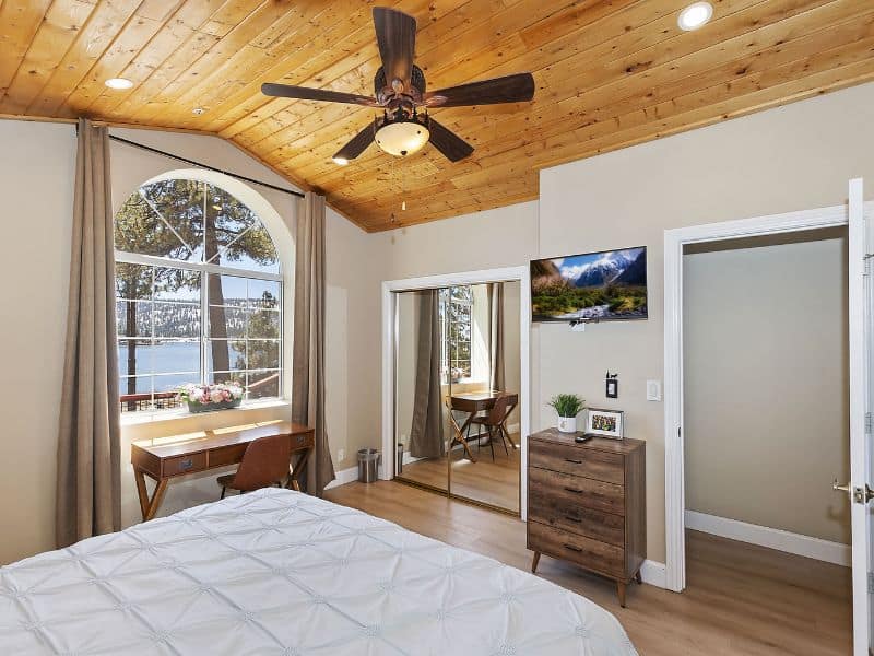Bedroom inside a Big Bear Vrbo rental, with a large window offering a view of the lake, a wooden ceiling fan, and minimalist decor.