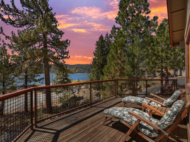 View from a Big Bear lakefront cabin rental deck featuring two lounging chairs overlooking the lake at dusk, framed by tall pine trees.