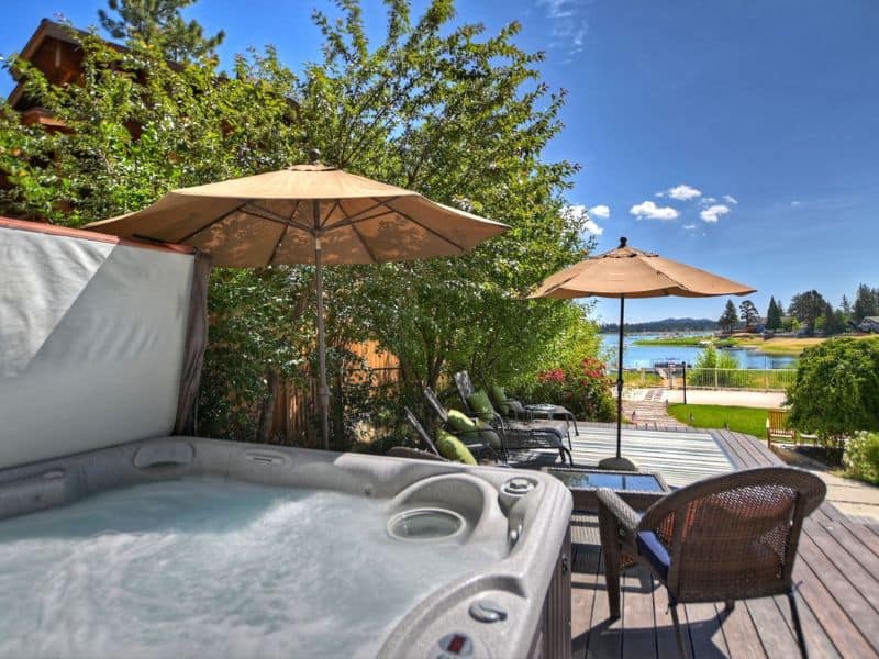 Deck with a hot tub and patio furniture shaded by umbrellas at a Big Bear lakefront cabin rental, with a clear view of the lake and green lawn.