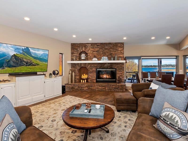 Comfortable living area with a large stone fireplace and flat-screen TV in a Big Bear rental, with ample seating and lake views through the balcony doors.