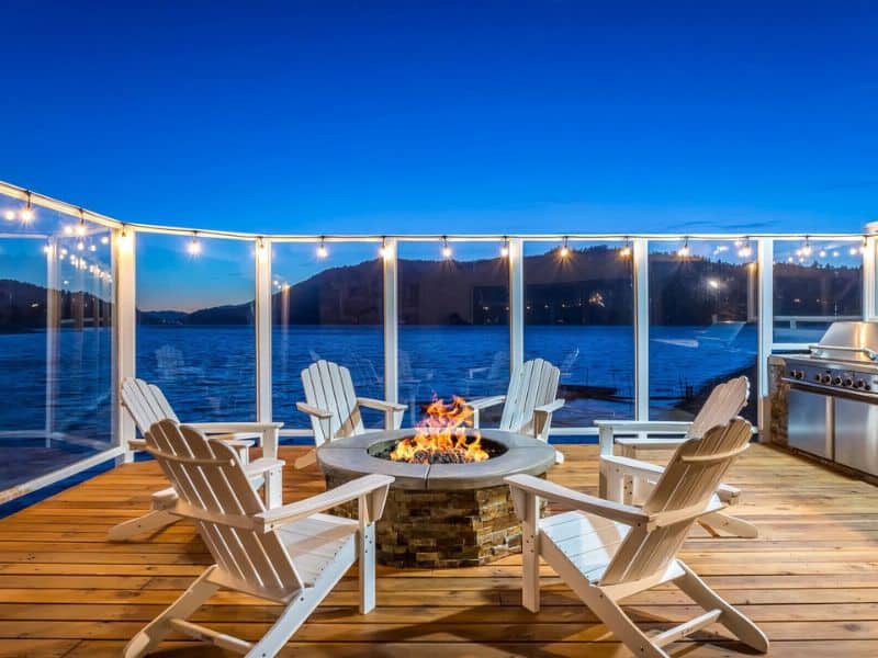 Evening view from a deck of a Big Bear Vrbo rental, featuring Adirondack chairs around a fire pit and string lights with the lake in the background.