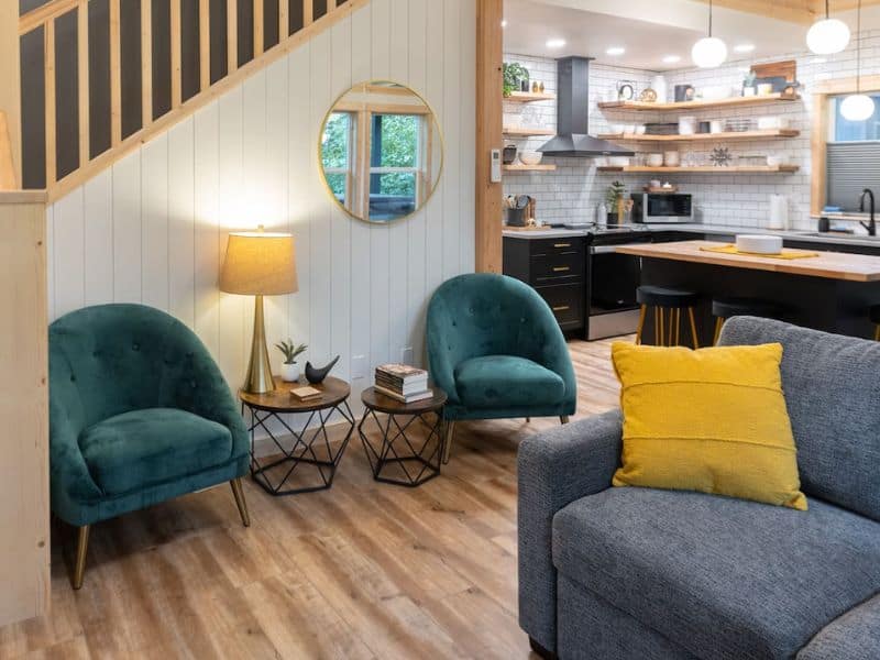 Interior shot of a cozy cabin near Mount Rainier featuring teal accent chairs, a gray sofa with a yellow cushion, and an open-concept kitchen with wooden shelves.