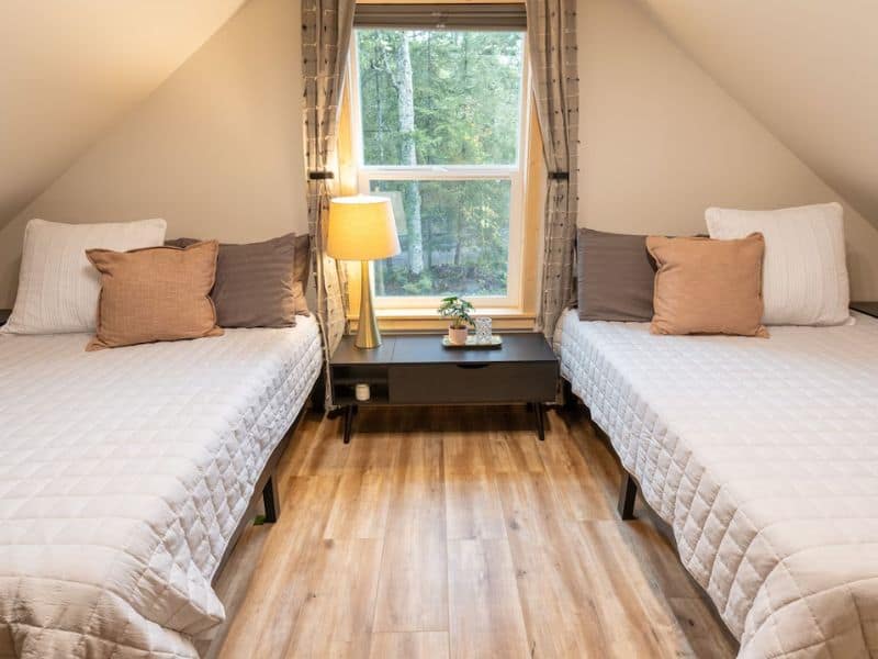Twin beds in a Mt Rainier cabin rental's upper-level bedroom with neutral bedding, a shared nightstand, and a window overlooking the woods.