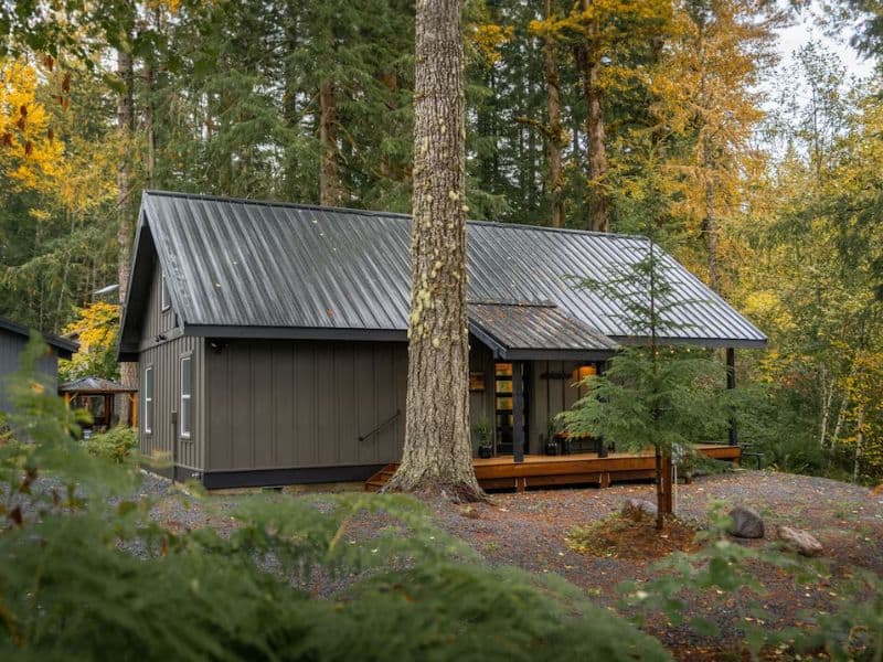 A secluded cabin surrounded by trees near Mount Rainier, with a distinctive A-frame design, gray siding, and a welcoming front porch with benches.