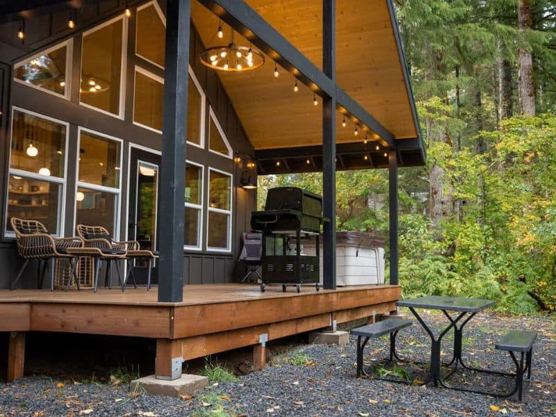 Exterior view of a modern Mount Rainier cabin with a spacious wooden deck, outdoor furniture, and a barbeque grill, nestled in a forest setting.