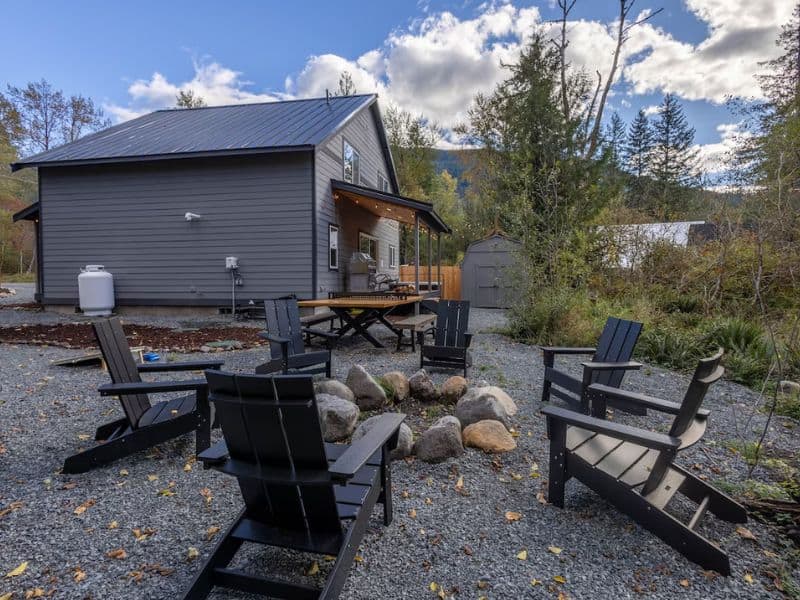 Outdoor firepit area of a Mount Rainier cabin with Adirondack chairs and picnic table, set against a backdrop of trees with mountain views peaking in the distance.
