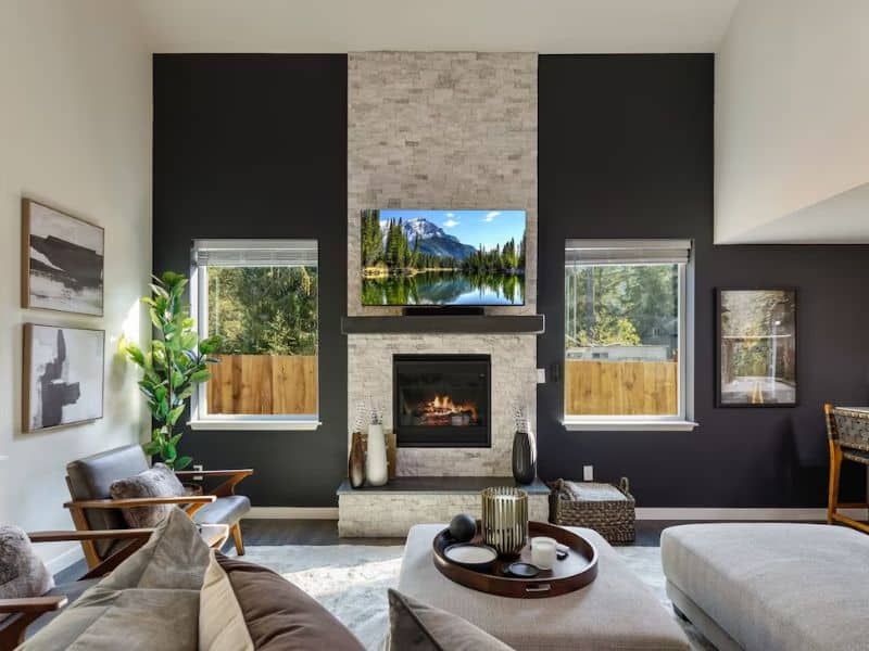 Modern living room inside a Mount Rainier cabin rental with high ceilings, stone fireplace, and windows framing the outdoors, complemented by contemporary furnishings and art.