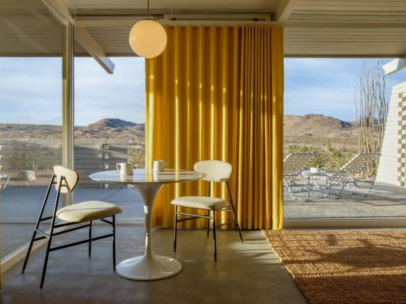 Inside a bright bungalow with floor-to-ceiling windows draped with vibrant yellow curtains, providing a cozy dining nook that captures the essence of Joshua Tree's natural beauty.