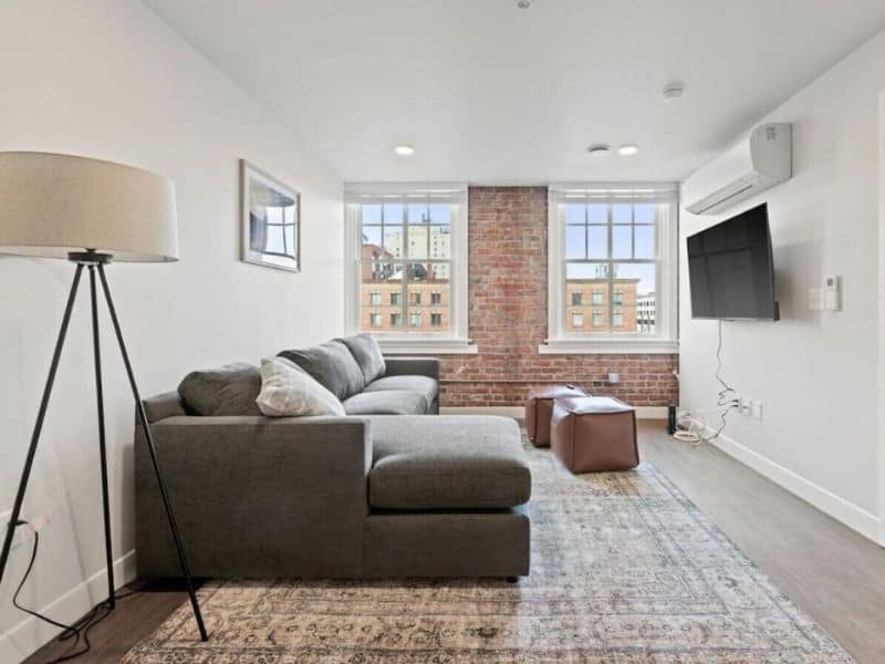 Interior of a modern 2-bedroom rental in Spokane with a sectional gray sofa, exposed brick wall, flat-screen TV, and large windows providing a view of the city.