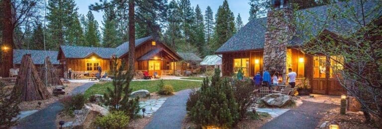 Evening view of Evergreen Lodge at Yosemite, featuring cozy wooden cabins with warm lights, nestled among tall pine trees. Guests can be seen gathering around an outdoor fireplace, enjoying the serene forest setting.