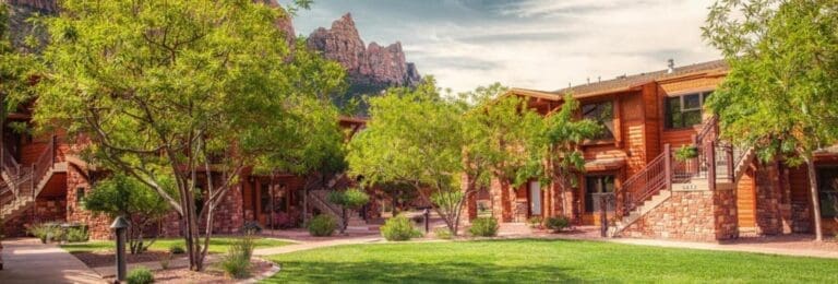 Cable Mountain Lodge in Zion National Park, showcasing wooden lodging surrounded by greenery and trees, with dramatic red rock cliffs in the background.