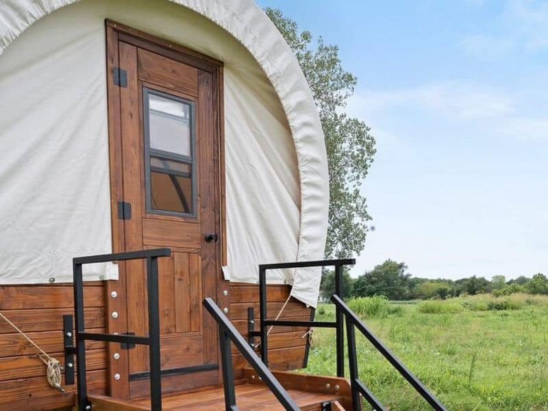 A close-up of the entrance to a covered wagon-style glamping tent with a wooden door and steps leading up to the entrance. The wagon is set in a grassy open field. Grand Canyon Glamping Resort.
