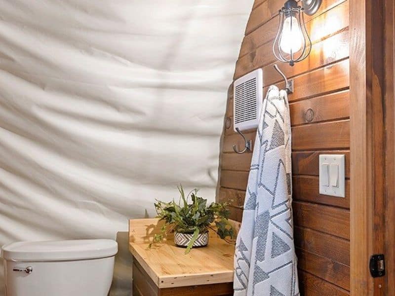 A small bathroom with a toilet, a wooden vanity with a potted plant, and a towel hanging from a rustic light fixture mounted on a wooden wall.