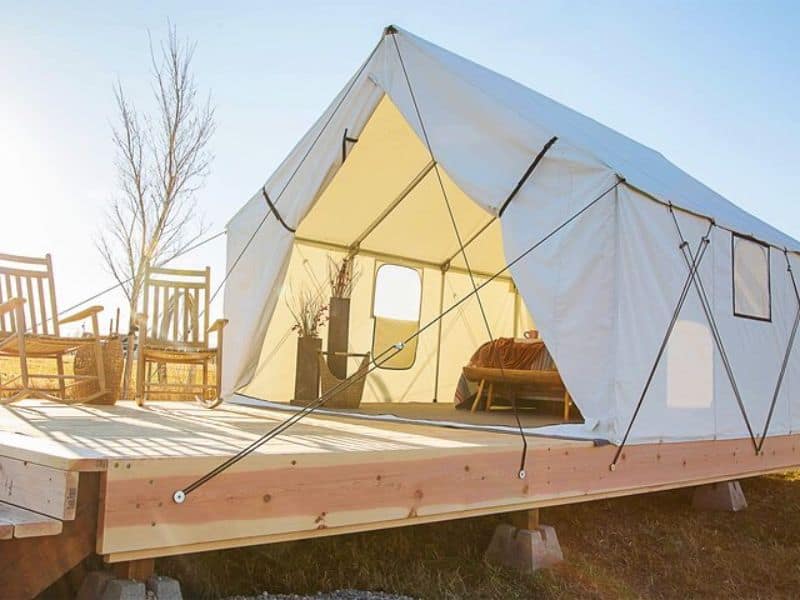 A glamping tent set on a wooden platform with two rocking chairs on the deck in front of the tent. The tent's flaps are open, revealing an interior with a well-made bed. Glamping tent at Grand Canyon Glamping Resort.