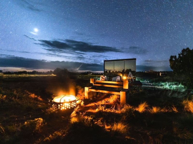 A nighttime view of the "Grand Canyon Stargazer" glamping site, with a fire pit and wooden bench in the foreground. The sky is filled with stars, and a small, mirrored cabin reflects the surrounding landscape.