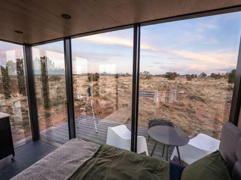 Interior view from "Grand Canyon Stargazer" cabin with panoramic floor-to-ceiling windows offering uninterrupted views of the desert landscape. A telescope is set up on the deck outside.