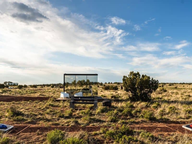 A view of "Grand Canyon Stargazer" cabin in an open desert landscape. The mirrored exterior of the cabin reflects the sky and surroundings, making it almost disappear into the environment.
