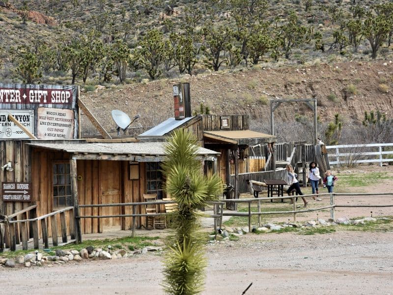 Visitors strolling by the wooden structures and signage of the Grand Canyon Western Ranch's cowboy-themed lodging in a rugged desert setting.