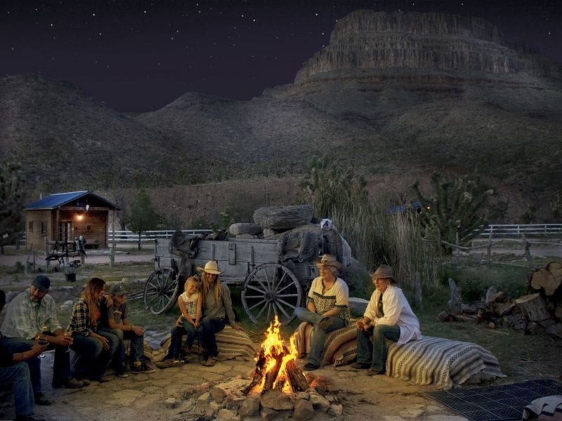 Group gathered around a campfire for a night under the stars at Grand Canyon Western Ranch, with a historic wagon and cabin in the background.