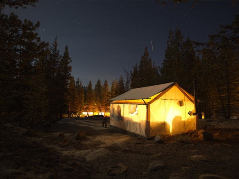 Illuminated canvas tent at night in the heart of Yosemite National Park, surrounded by the forest under a starry sky.