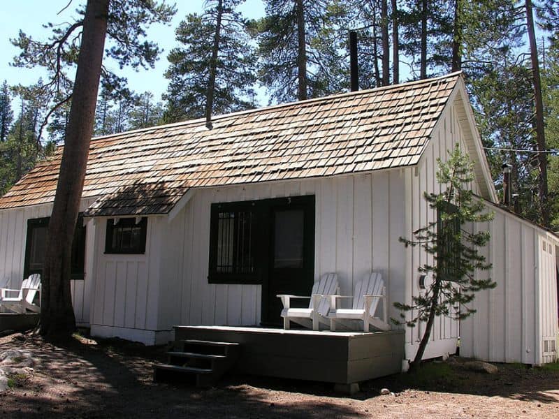 A white cabin with a sloped shingle roof and a small porch with two white Adirondack chairs, among tall pines at White Wolf Lodge in Yosemite National Park.