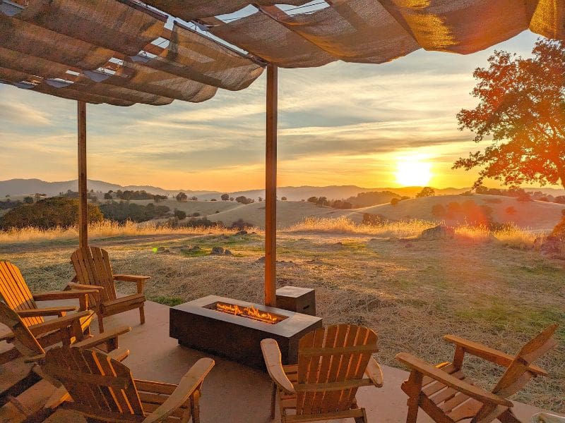 Sunset view from a cozy glamping site at Wildhaven Yosemite, with a fire pit and chairs overlooking the rolling hills.