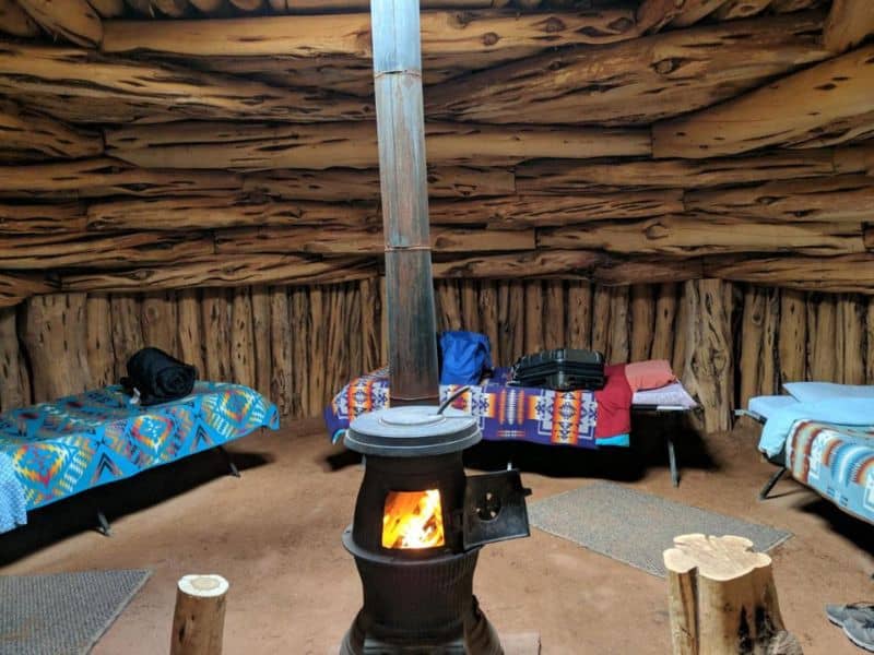 Rustic interior of a traditional Navajo hogan with log walls, multiple beds covered with colorful blankets, and a central wood stove providing warmth.