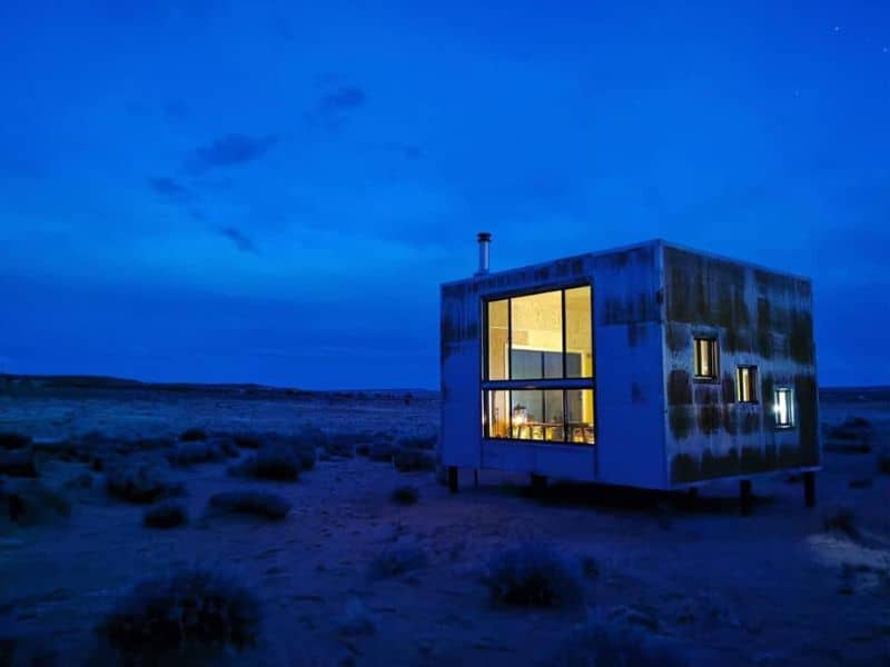 A modern, cube-shaped glamping structure illuminated from within stands out against the twilight sky in the Grand Canyon area, offering a unique Navajo lodging experience.