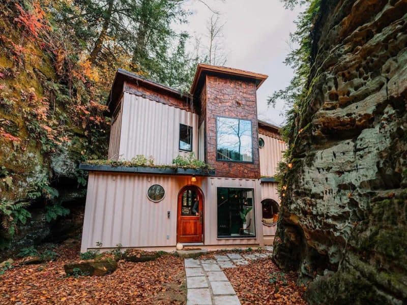 Exterior view of The Roca Box Hop, a distinctive Hocking Hills container cabin, nestled among the trees with an autumnal backdrop and a rugged stone pathway leading to its entrance.