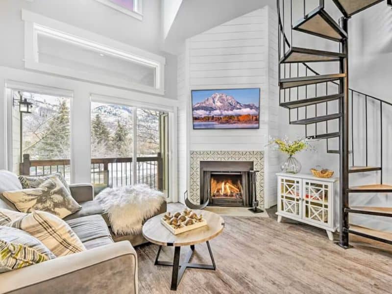 Interior of a two-bedroom condo rental in Park City, Utah, featuring a living area with a lit fireplace, a spiral staircase, and a television displaying a mountain image. Large windows offer a view of the outside landscape.